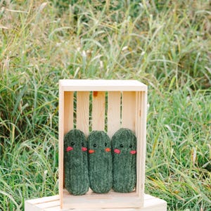 May include: Three green plush pickle toys with embroidered faces are arranged in a wooden crate. The crate is sitting on a wooden box.