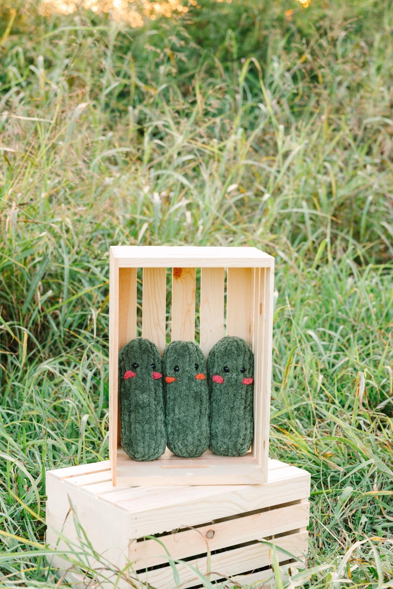 May include: Three green plush pickle toys with embroidered faces are arranged in a wooden crate. The crate is sitting on a wooden box.