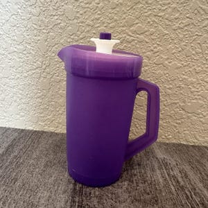 May include: A purple plastic pitcher featuring a matching purple lid with a white center insert and a small purple knob on top. The pitcher has a sturdy side handle and a molded pouring spout, designed with a simple, functional cylindrical body.