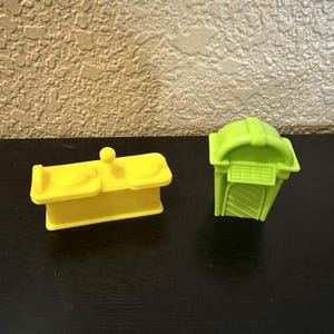 May include: Two plastic toy structures. One is yellow and rectangular with two circular features and a small ball on top. The other is lime green, with a curved roof and a slatted front.