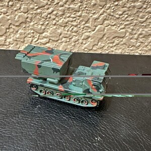 May include: A miniature toy military vehicle featuring a tracked chassis and a large rectangular launcher box on top. The vehicle is painted in a camouflage pattern consisting of olive green, black, and bright orange patches across its entire body.
