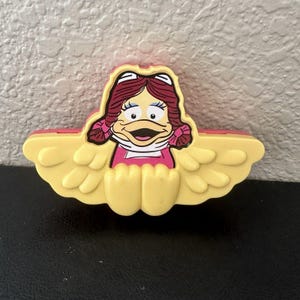 May include: A yellow and pink plastic toy featuring a cartoon duck character with red hair and a pink dress. The duck is smiling with its mouth open. The toy has yellow wings and a pink border.