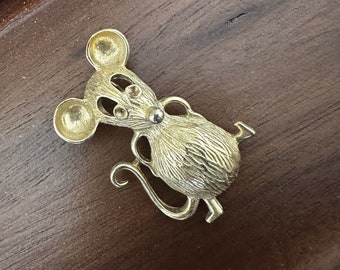 May include: A gold-toned metal brooch in the shape of a cartoon mouse. The mouse has large ears, a long tail, and a textured body.
