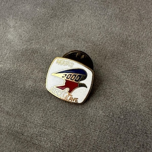 May include: A square-shaped metal lapel pin with rounded corners, featuring a white enamel background. The design includes a stylized eagle graphic in blue and red, with the number 2000 in the center. The text HOURS is above and SICK LEAVE is below.