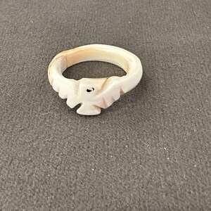May include: A carved ring featuring a stylized bird design. The ring is made of a light-colored material, possibly bone or a similar substance. The bird is depicted with outstretched wings and a small, dark eye.