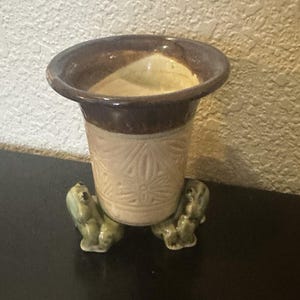 May include: A ceramic vase with a brown rim and a beige body, supported by three green frog figurines. The vase has a floral pattern and is suitable for holding small plants or flowers.