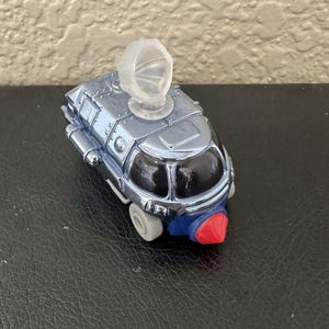 May include: A metallic blue toy vehicle with a clear plastic dome on top. The toy has a red nose, blue accents, and small gray wheels. The vehicle's design suggests a futuristic or space-themed concept.