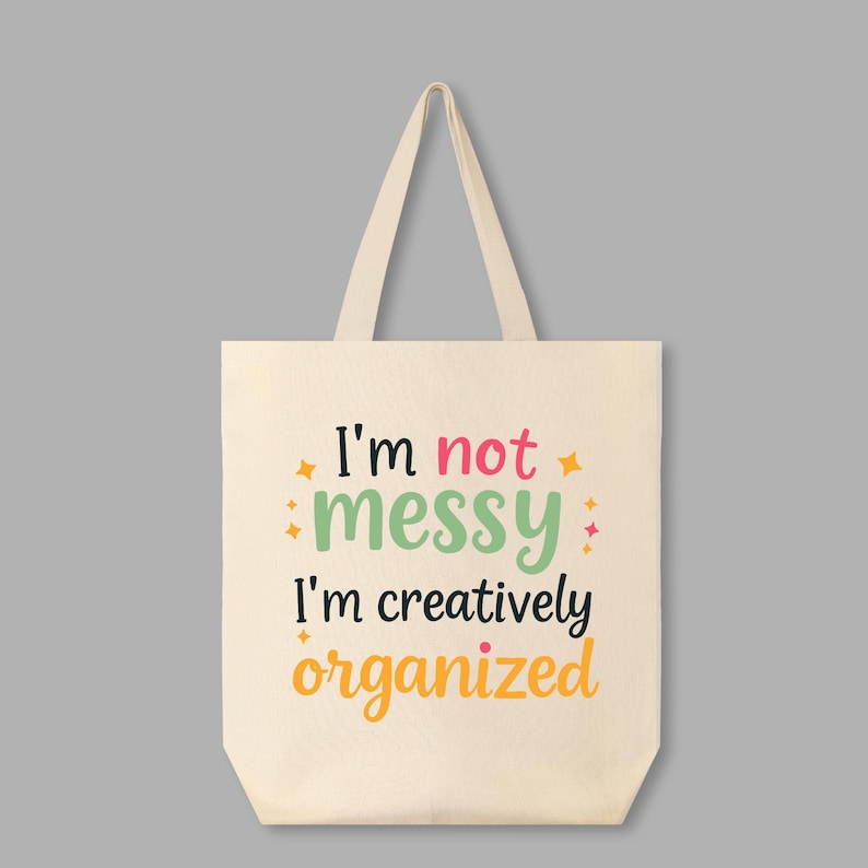 May include: A white canvas tote bag with the text "I'm not messy I'm creatively organized" in black, green, pink, and orange lettering. The text is surrounded by small yellow stars.
