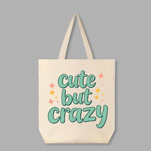 May include: A white canvas tote bag with the text "cute but crazy" in teal green lettering with pink and yellow stars.