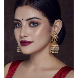 May include: A pair of gold earrings with white pearl accents. The earrings are in the shape of a bell with a floral design at the top. The earrings are worn by a person with dark hair and a red blouse.