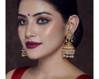 May include: A pair of gold earrings with white pearl accents. The earrings are in the shape of a bell with a floral design at the top. The earrings are worn by a person with dark hair and a red blouse.