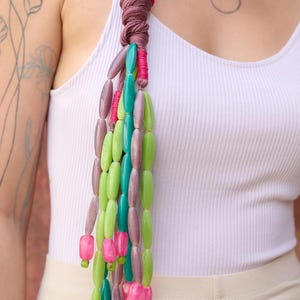 May include: A white tank top with a braid in the hair, decorated with a long strand of colorful beads. The beads are green, pink, and purple.