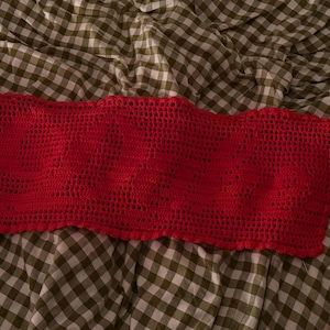 May include: A vibrant red, handmade crochet top with a scalloped edge. The open weave design is visible, and the top is laid on a green and white checkered fabric. The top is a stylish piece of clothing.