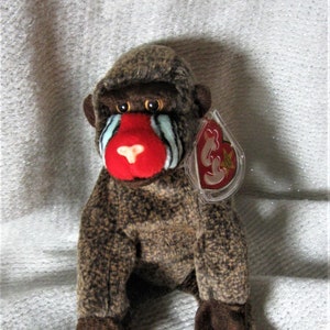 May include: A plush mandrill toy with brown mottled fur, a bright red nose, and light blue cheek markings. The stuffed animal features dark brown hands and feet, amber-colored eyes, and a heart-shaped plastic tag attached to the ear with the Ty logo.