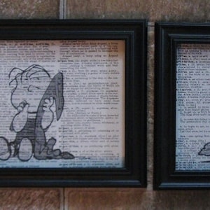 May include: Two black framed prints of Peanuts characters on a white background with black text. The print on the left shows Charlie Brown holding a blanket. The print on the right shows Linus holding a blanket.
