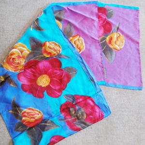 May include: A rectangular scarf featuring a vibrant floral pattern of large red, orange, and yellow camellia flowers with dark green leaves. The fabric is divided into two distinct sections, one with a bright turquoise blue background and one with a purple background.