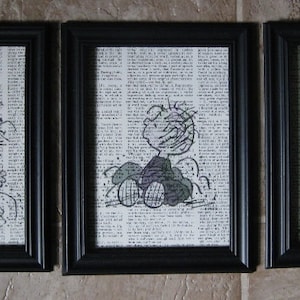 May include: Three black framed prints of a cartoon character with different expressions. The character is drawn in black and white on a background of printed text.