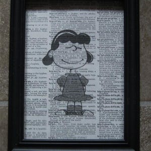 May include: Framed black and white print featuring a cartoon girl with pigtails and glasses, set against a dictionary page background. The artwork is a whimsical illustration.