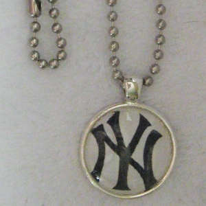 May include: A silver-colored ball chain necklace with a round pendant. The pendant features the black and white New York Yankees logo. The logo is encased in a clear, circular setting.