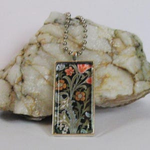 May include: A rectangular pendant necklace featuring a black background with a detailed floral pattern in shades of orange, green, and white. The pendant is encased in a thin silver-toned metal frame and hangs from a silver-toned ball chain.
