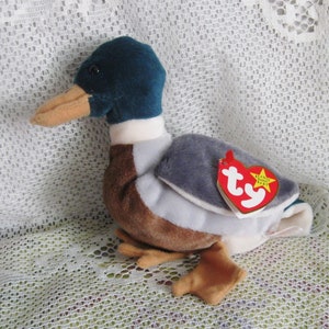 May include: A teal, brown, and gray plush duck with a white collar. The duck has a Ty tag attached to its wing that says "Beanie Babies".