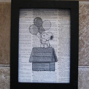 May include: Black framed print featuring Snoopy from Peanuts holding balloons, perched atop his doghouse. The artwork is set against a backdrop of dictionary pages, creating a vintage aesthetic.