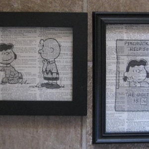 May include: Two framed art prints featuring Peanuts characters. One print shows Lucy and Charlie Brown, the other shows Lucy with the text "Psychiatric Help 5¢" and "The Doctor Is In". The prints are set against a background of vintage newspaper text.