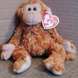 May include: A plush monkey toy with shaggy brown fur, a light tan face, and dark eyes. The toy features a heart-shaped red and white tag attached to its ear that reads Ty Original Beanie Babies. It has light tan hands and feet.