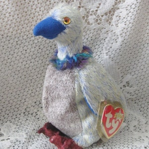 May include: A plush toy duck with a blue beak and feet, and a blue and purple collar. The duck has a white and blue body with a gray chest. A red heart-shaped tag is attached to the duck.