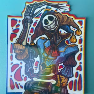 May include: A die-cut sticker featuring a stylized jester character with a skeletal arm, a wide grinning mask, and a blue and brown outfit. The design includes a vibrant red, blue, and yellow color palette with a distinct chromatic aberration effect.