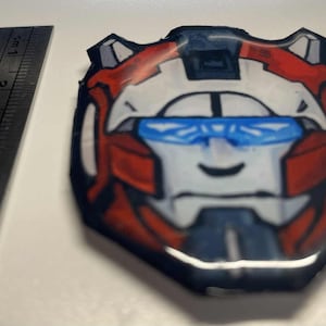 May include: A cartoon shield in red, white, and blue, with a smiling face and blue visor. A metal ruler with inch measurements is next to the shield. The shield is approximately 7 cm in length.