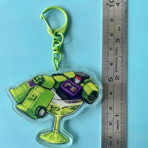 May include: A lime green acrylic keyring featuring a robot character lounging inside a martini glass filled with a green liquid. The keyring is attached to a lime green plastic clasp and is positioned next to a metal ruler measuring 10 centimetres.