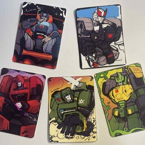 Puede incluir: Cinco tarjetas coleccionables ilustradas con personajes robot en varias poses. Las tarjetas muestran una gama de colores, incluyendo rojo, verde, gris y azul. Cada tarjeta tiene un diseño único, algunas con un fondo espacial.