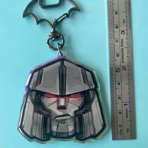May include: An acrylic keyring featuring a grey robot head with red eyes, attached to a black metal bat-shaped clasp. A stainless steel ruler is placed alongside the keyring, showing the robot head measures approximately 6.5 cm in height.