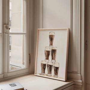 May include: A framed print featuring a watercolor illustration of six glasses of dark beer stacked in a pyramid formation. The artwork has a light wooden frame and is displayed on a windowsill next to a stack of books.