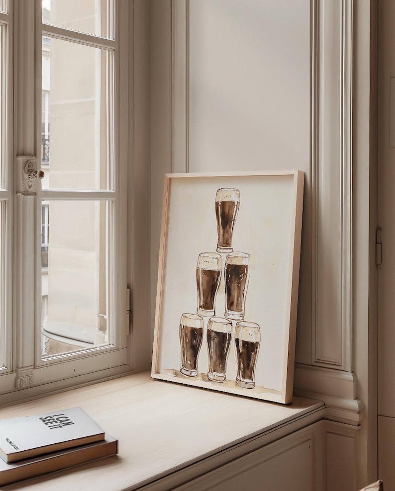 May include: A framed print featuring a watercolor illustration of six glasses of dark beer stacked in a pyramid formation. The artwork has a light wooden frame and is displayed on a windowsill next to a stack of books.