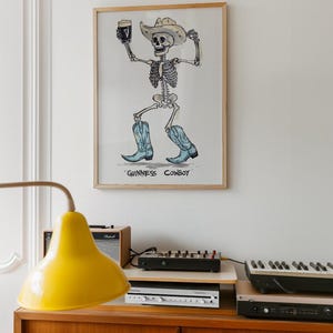 May include: A framed art print featuring a skeleton cowboy holding a pint of Guinness. The skeleton wears a cowboy hat and blue cowboy boots. The text "GUINNESS COWBOY" is below the image. The artwork is displayed above a wooden cabinet with electronic equipment.