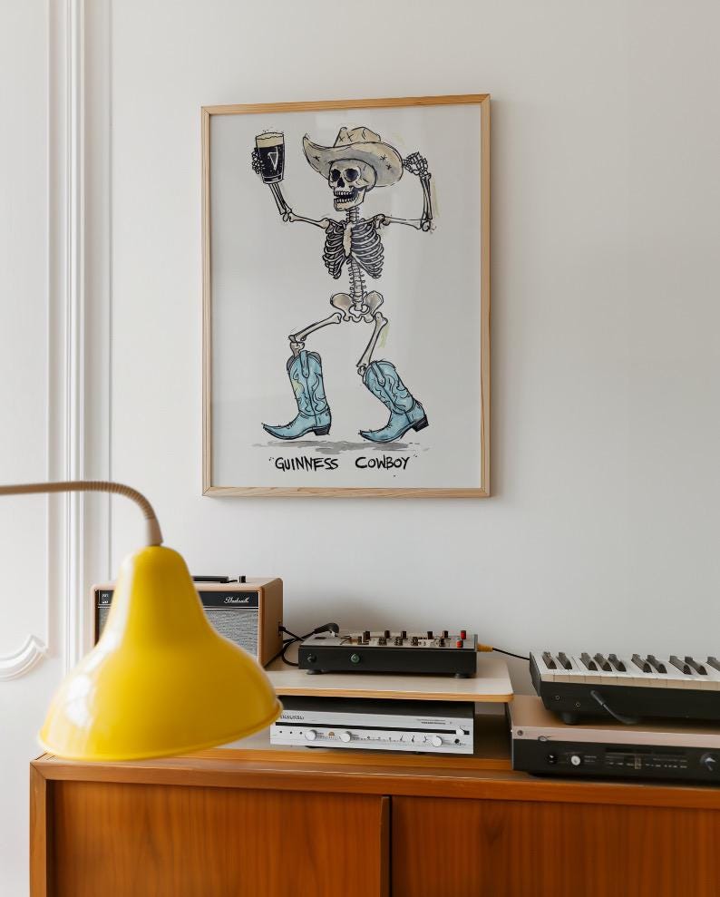 May include: A framed art print featuring a skeleton cowboy holding a pint of Guinness. The skeleton wears a cowboy hat and blue cowboy boots. The text "GUINNESS COWBOY" is below the image. The artwork is displayed above a wooden cabinet with electronic equipment.