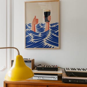 May include: A framed art print depicts two hands emerging from blue waves, one giving a thumbs-up and the other holding a pint of dark beer. The artwork has a light beige background and a wooden frame. The print hangs above a wooden cabinet with electronic equipment.