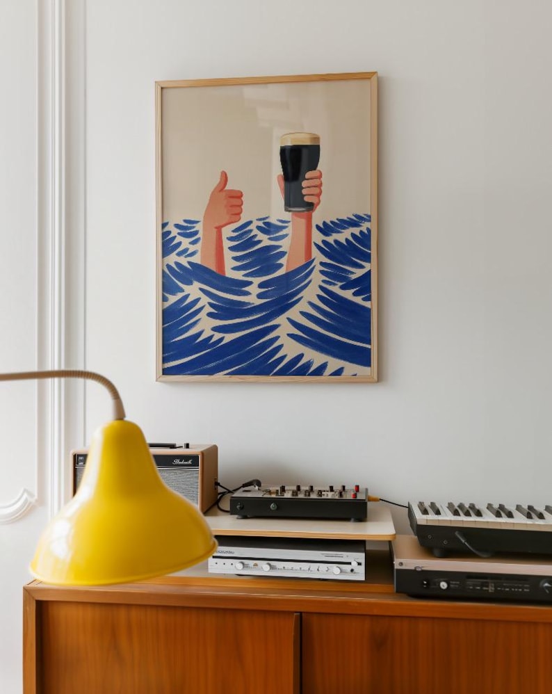 May include: A framed art print depicts two hands emerging from blue waves, one giving a thumbs-up and the other holding a pint of dark beer. The artwork has a light beige background and a wooden frame. The print hangs above a wooden cabinet with electronic equipment.