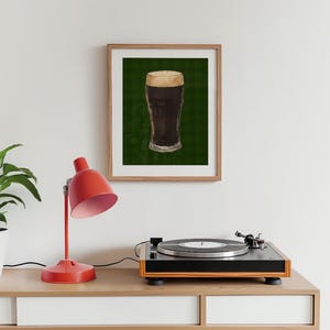 May include: A framed print depicting a pint of dark beer with a creamy head. The image is set against a green, grid-patterned background. The print is in a wooden frame, displayed on a white surface with a red desk lamp and a record player.