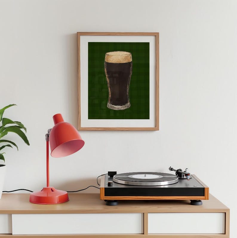 May include: A framed print depicting a pint of dark beer with a creamy head. The image is set against a green, grid-patterned background. The print is in a wooden frame, displayed on a white surface with a red desk lamp and a record player.