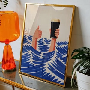 Op de afbeelding: Kunstprint in een gouden lijst met handen die een pint donker bier boven blauw water vasthouden, met een duim omhoog. Het kunstwerk heeft een witte achtergrond met een zon. Op een tafel.