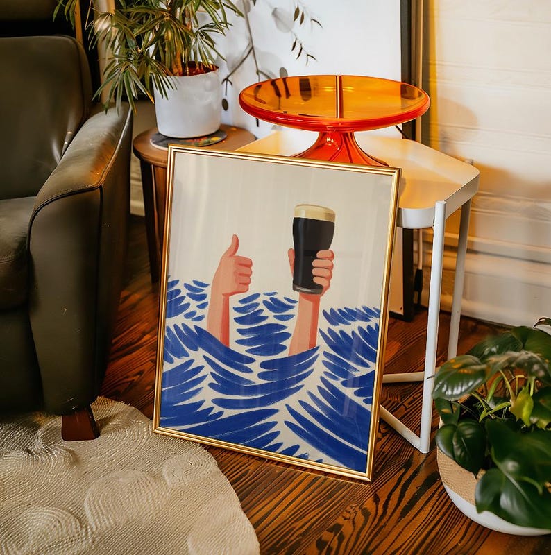 May include: A framed art print with a gold frame. The artwork depicts two arms emerging from blue waves, one giving a thumbs-up and the other holding a pint of dark beer. The background is a light cream color.