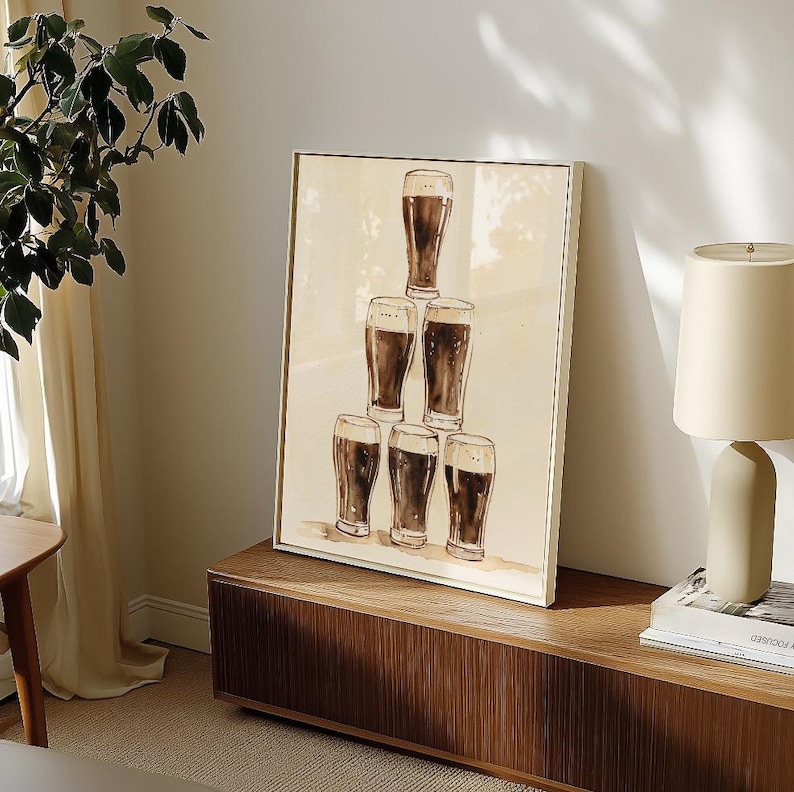May include: A framed print featuring a pyramid of seven beer glasses filled with dark liquid. The artwork has a neutral background and is displayed on a wooden cabinet. The print has a light-colored frame.
