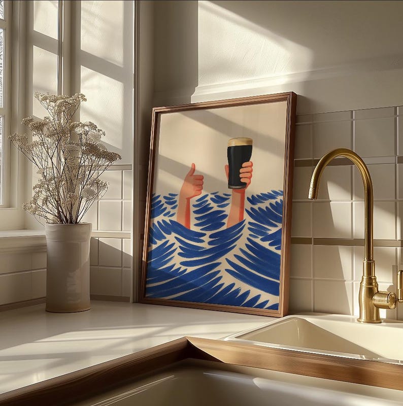 May include: Framed art print featuring a whimsical illustration of hands holding a pint of dark beer above blue waves. The artwork has a light beige background and is set in a wooden frame. The image is displayed on a white countertop with a gold faucet.