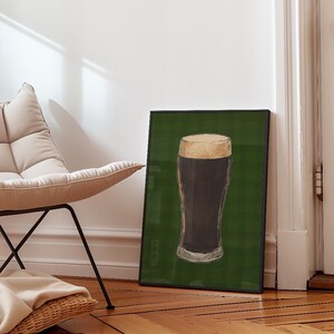 May include: A framed print depicting a pint of dark beer with a creamy head. The artwork is set against a green plaid background. The print is displayed on a wooden floor next to a chair.