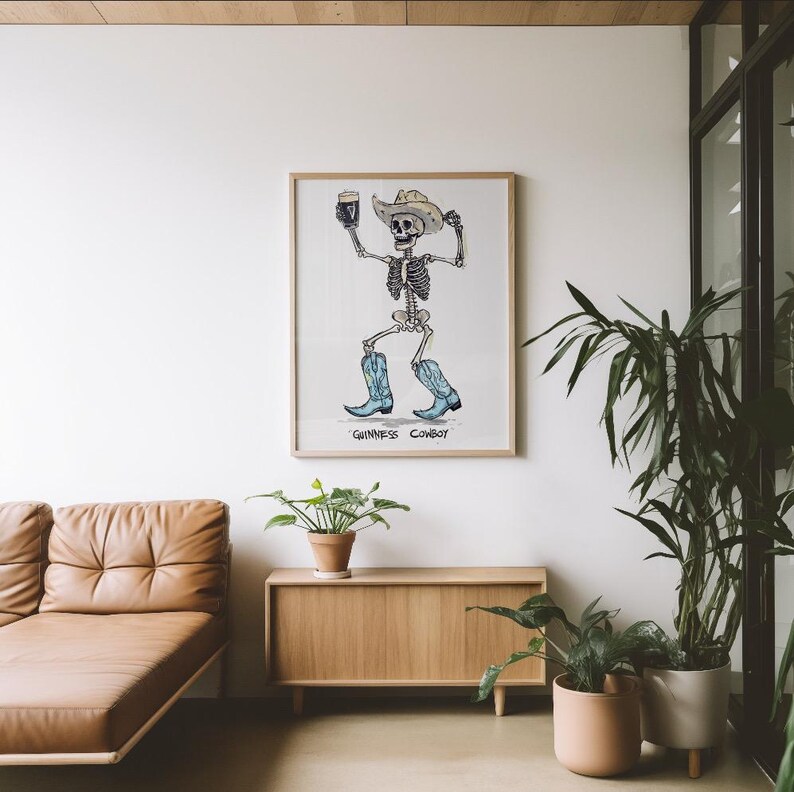 May include: A framed print featuring a skeleton cowboy holding a pint of beer. The skeleton wears a cowboy hat and blue boots. The text "Guinness Cowboy" is at the bottom of the print. The artwork is displayed on a white wall.