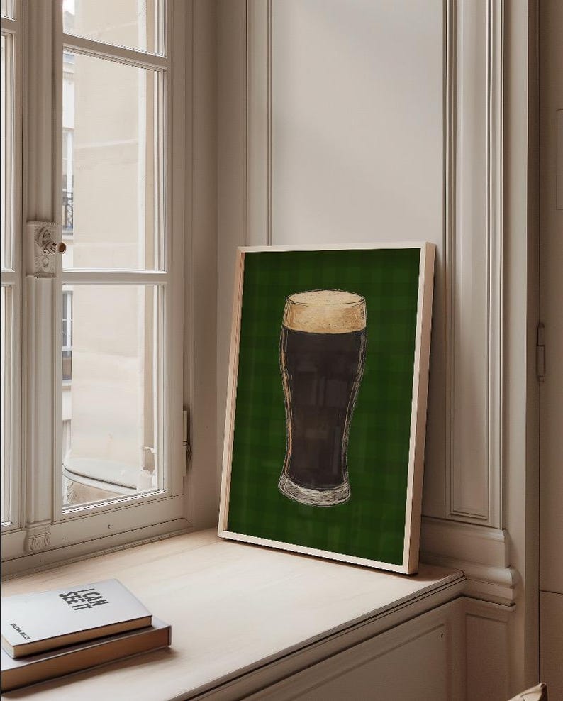 May include: A framed print depicting a pint of dark beer with a creamy head, set against a green plaid backdrop. The artwork is displayed on a windowsill next to a window and a stack of books.