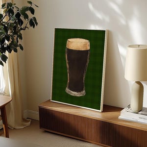 May include: A framed print depicting a pint of dark beer with a foamy head. The artwork is set against a green plaid background. The print is displayed on a wooden cabinet.